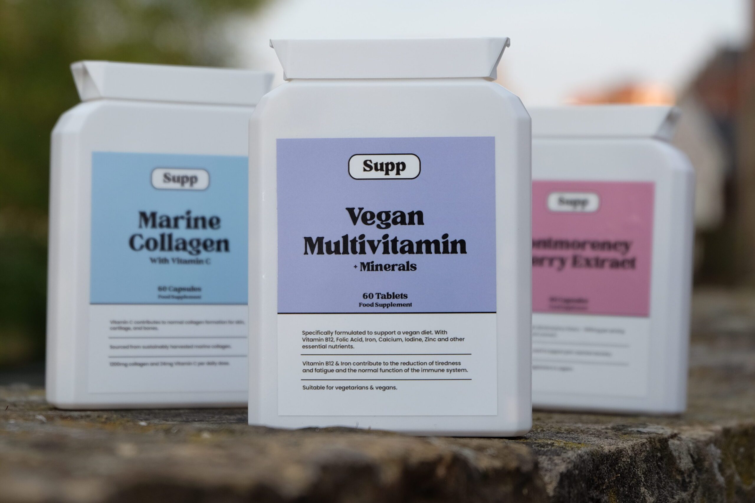 Three Supp supplement bottles on an outdoor stone surface, featuring Vegan Multivitamin in focus, with Marine Collagen and Montmorency Cherry Extract in the background.