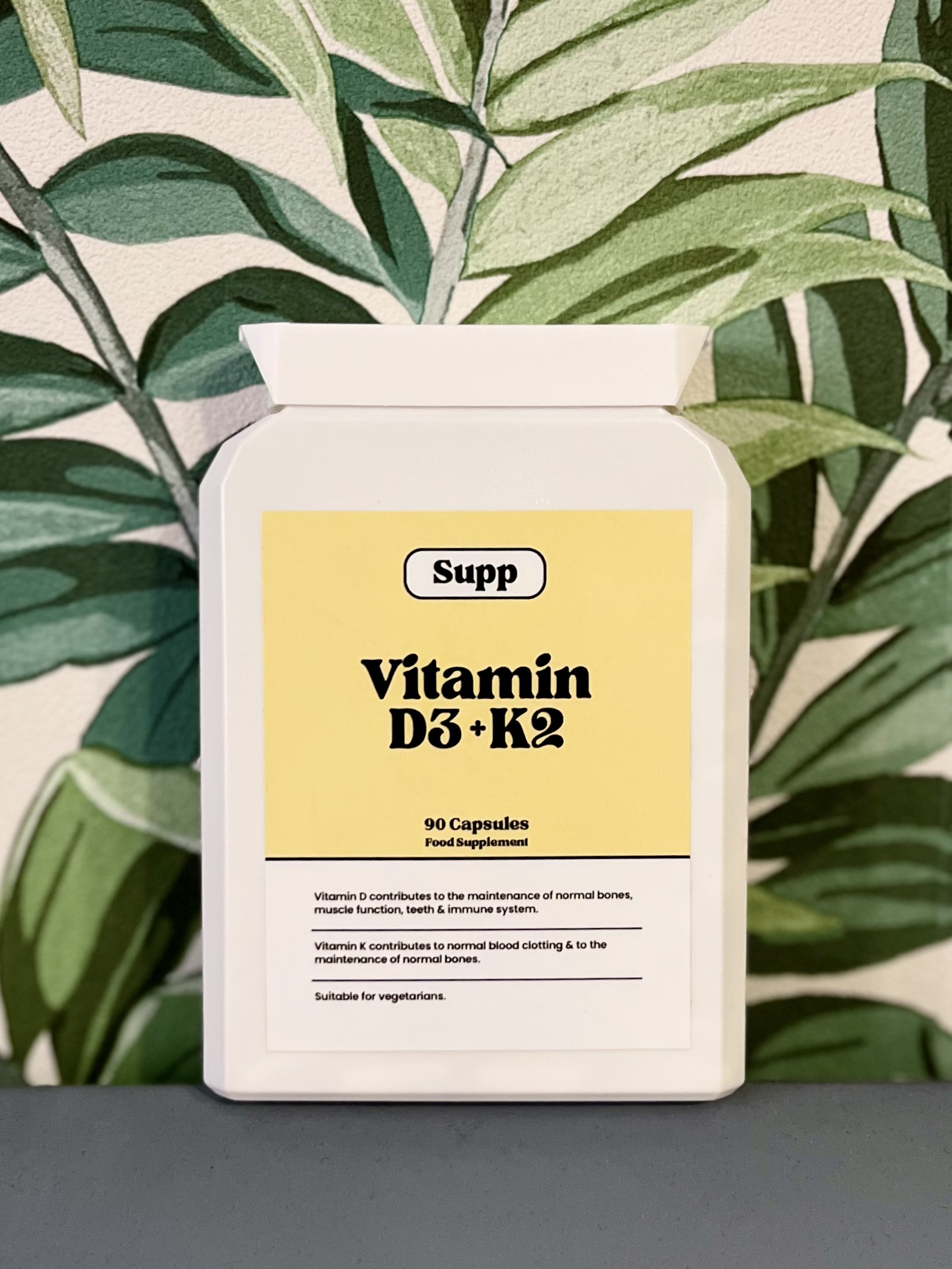 Supp Vitamin D3 & K2 bottle with a tropical plant wallpaper background.