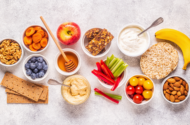 Selection of healthy snacks on a light background, including nuts, dried fruit, blueberries, apple, banana, yogurt, hummus, rice cakes, crackers, and vegetable sticks.