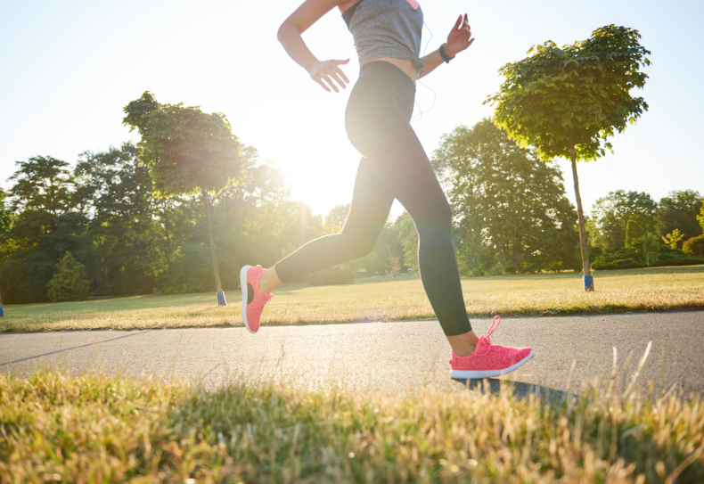 Woman jogging outdoors in a sunlit park, wearing black leggings, a grey sports top, and bright pink running shoes, with trees and greenery in the background