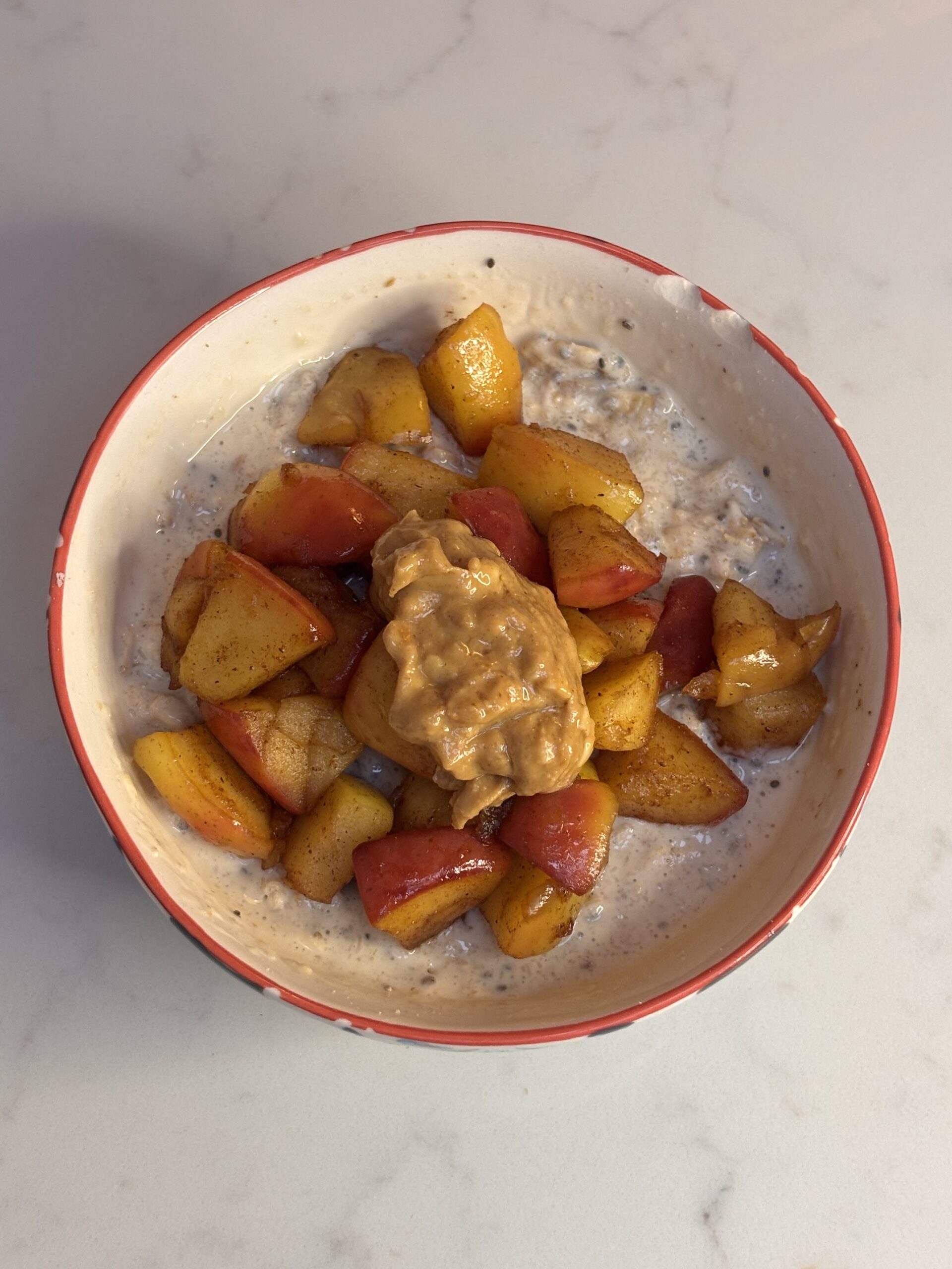 Bowl of apple cinnamon protein oats topped with warm cinnamon-spiced apple pieces and a spoon of crunchy peanut butter.