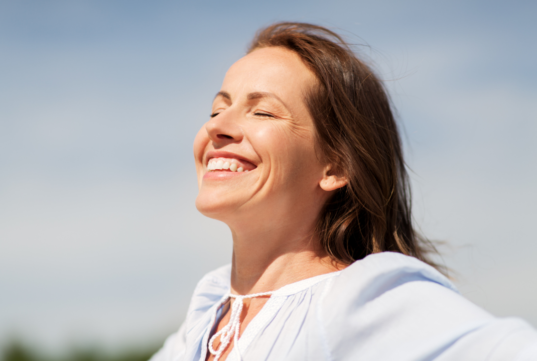 Woman taking a deep breath outdoors, smiling and soaking up the sunshine, looking relaxed.