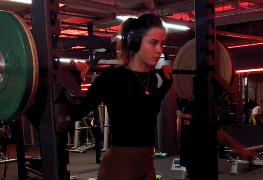 A woman in a gym performing a barbell squat on a rack, wearing headphones and a black long sleeve top, with red lighting in the background.