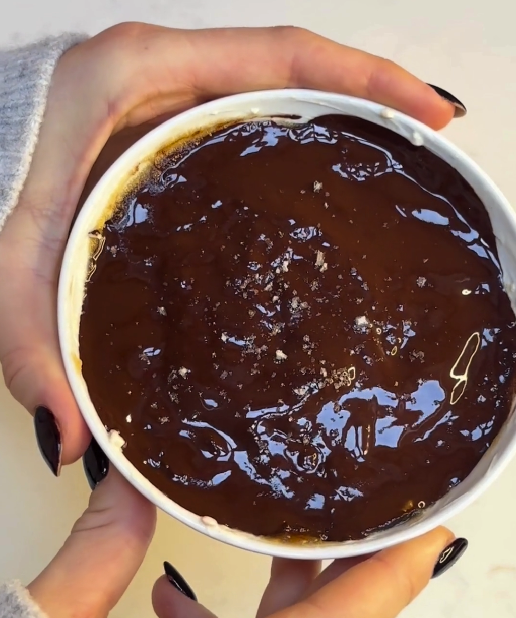 Hands holding a bowl of Millionaire Shortbread Overnight Oats topped with a glossy layer of melted dark chocolate and a light sprinkle of sea salt.