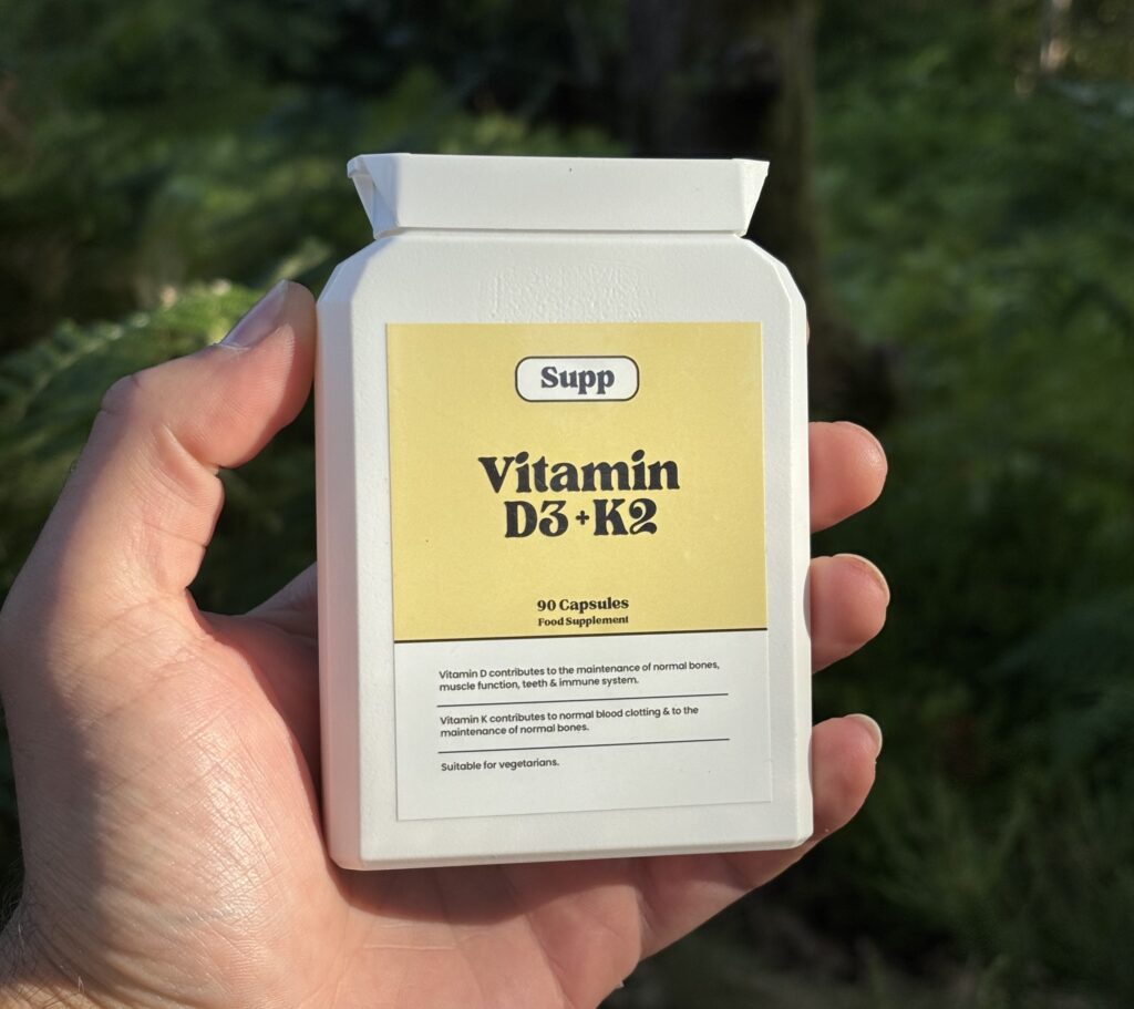 Hand holding Vitamin D3 + K2 supplement bottle in bright sun lighting with greenery in background