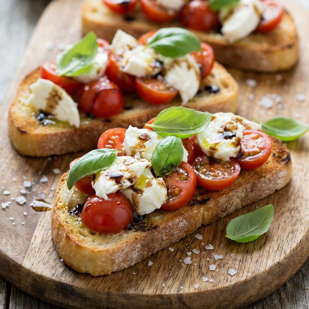 Caprese-Bruschetta-Bites Close-up of toasted ciabatta slices topped with juicy halved cherry tomatoes, torn fresh mozzarella and bright basil leaves, drizzled with olive oil and balsamic glaze, arranged on a wooden board with a scattering of sea salt
