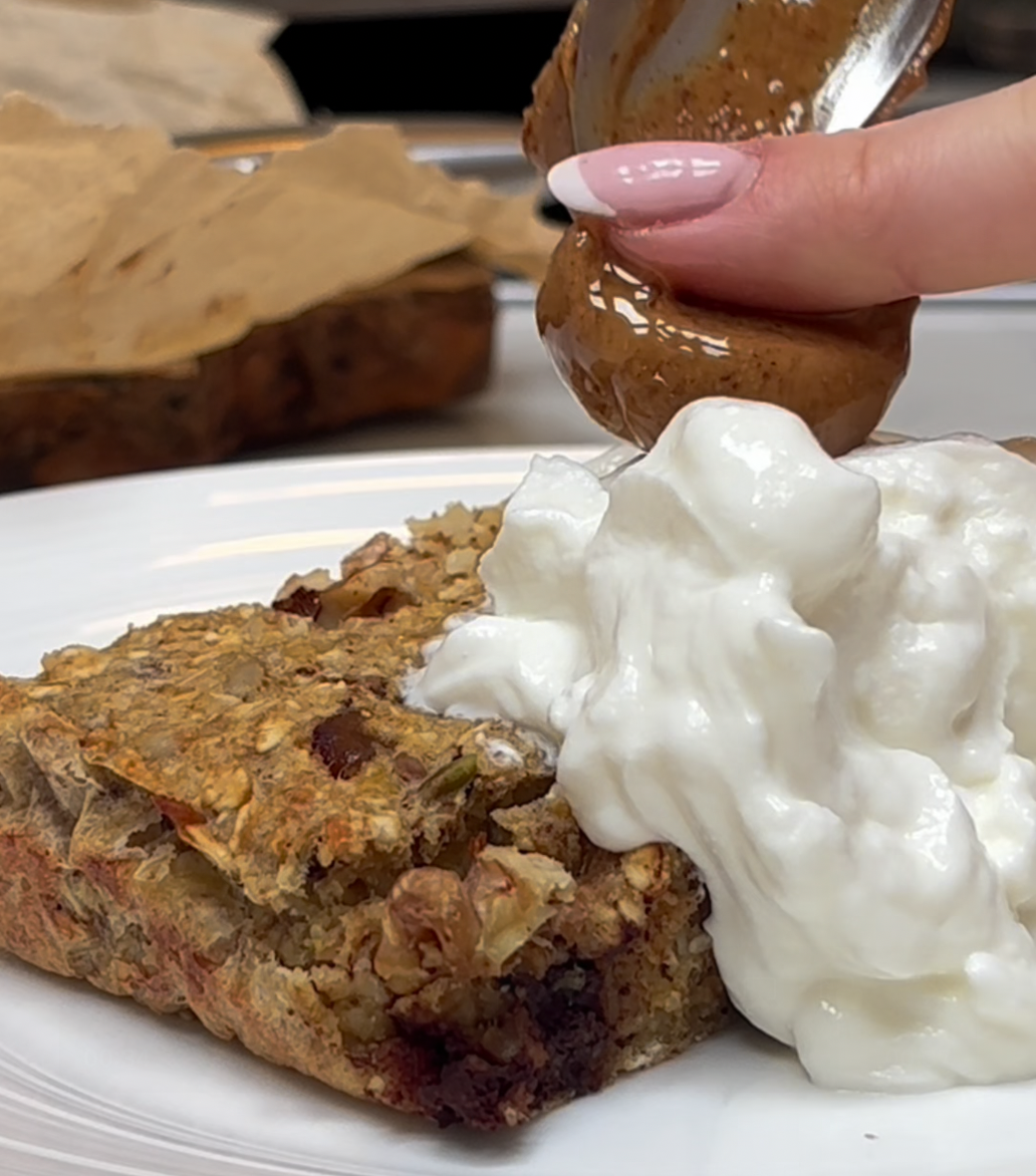 Banana and date baked oat square served with Greek yoghurt, topped with nut butter on a white plate.